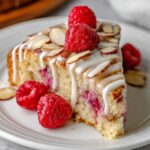 A slice of Raspberry Almond Cake drizzled with icing, topped with fresh raspberries and slivered almonds, showcasing a moist interior with vibrant raspberry filling.