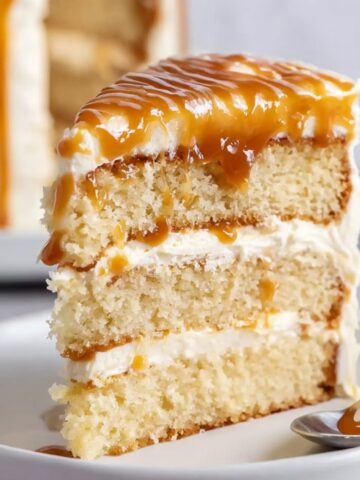 Easy Homemade Salted Caramel Cake 1 A slice of moist Salted Caramel Cake with layers of smooth buttercream frosting, topped with a rich drizzle of salted caramel sauce. The cake is served on a white plate with a spoon beside it.