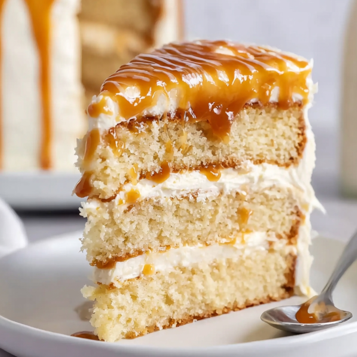 A slice of moist Salted Caramel Cake with layers of smooth buttercream frosting, topped with a rich drizzle of salted caramel sauce. The cake is served on a white plate with a spoon beside it.