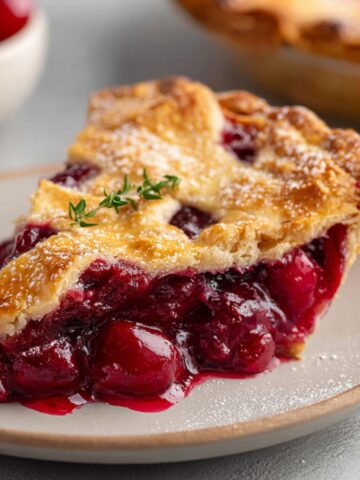 A slice of Sour Cherry Pie served on a plate, with a lattice crust, fresh cherries, and a sprinkle of powdered sugar, garnished with a sprig of thyme.