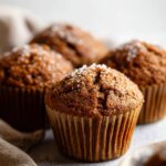 A close-up of freshly baked Spiced Molasses Gingerbread Muffins in paper liners, with a sprinkle of sugar on top, placed on a clean, soft background.