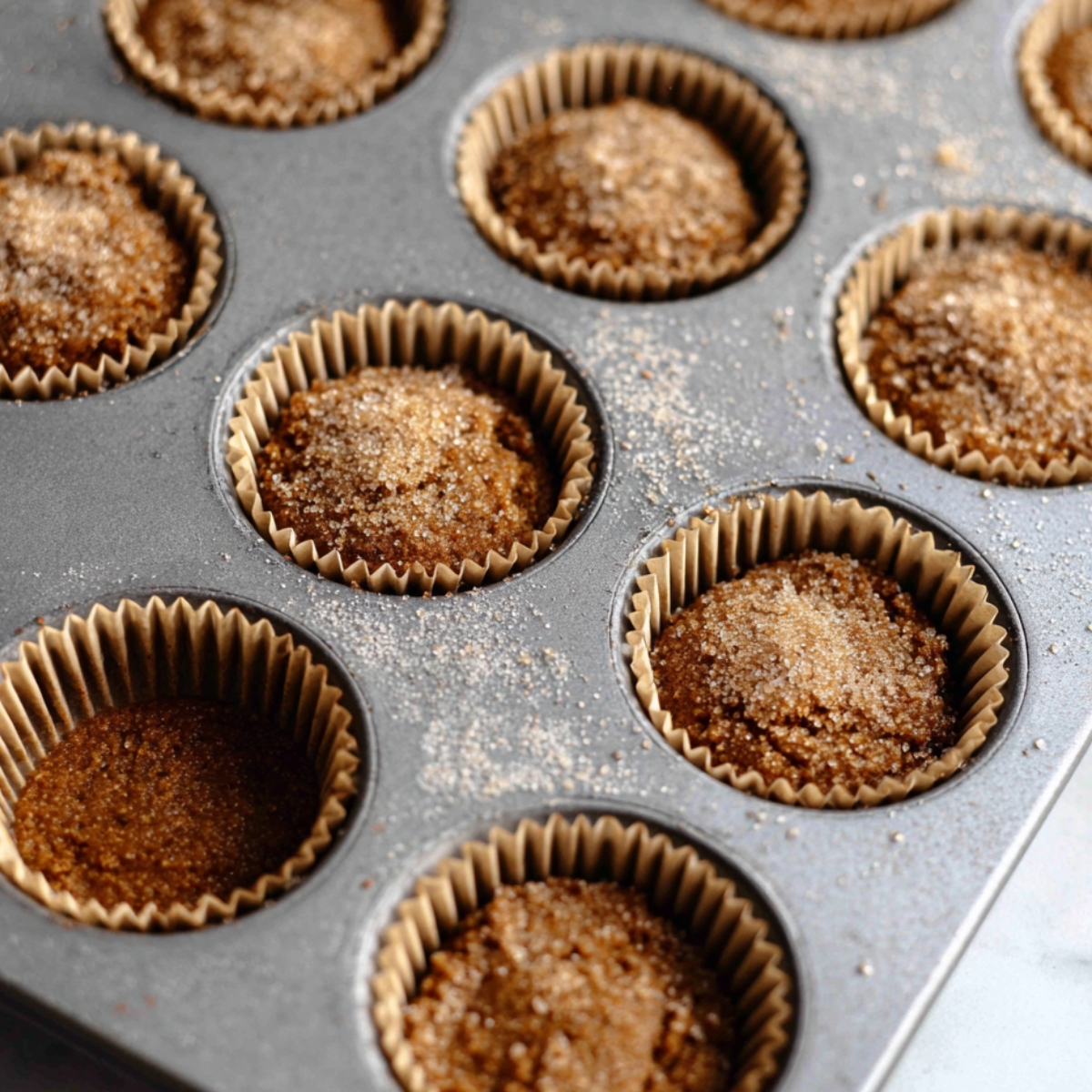 Spiced Molasses Gingerbread Muffins 11 A muffin tin filled with freshly baked spiced molasses gingerbread muffins, topped with a sprinkle of sugar, placed on a light-colored surface.