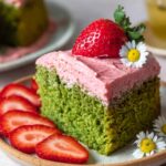A slice of Strawberry Matcha Latte Cake with strawberry frosting, topped with a fresh strawberry and a daisy, served with sliced strawberries.