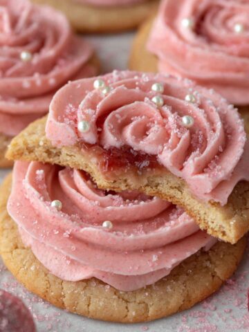 Delicious Sugar Plum Fairy Cookies 4 A Sugar Plum Fairy Cookies topped with pink buttercream frosting piped into a rosette shape, decorated with edible pearls and a sprinkling of sugar, with a bite taken revealing the jam filling.