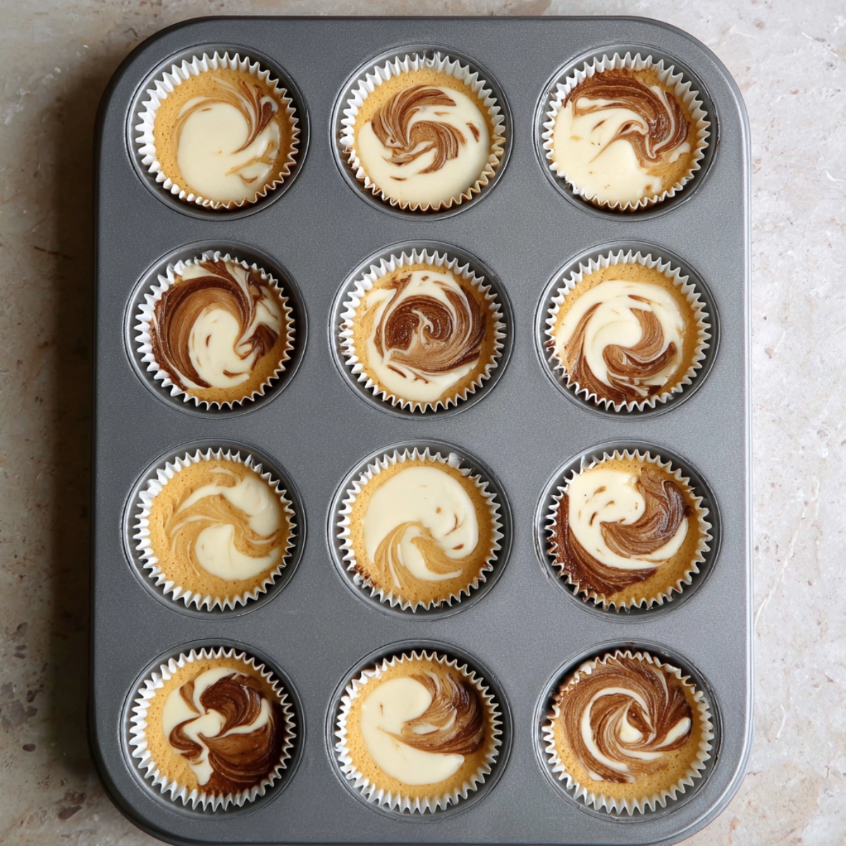 A muffin tin filled with tiramisu cupcake batter, with swirls of light and dark batters blended together, ready for baking.