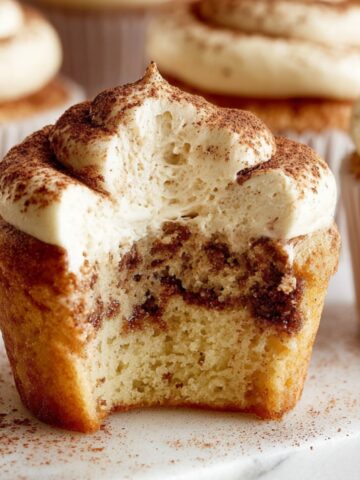 A close-up of a Tiramisu Cupcakes with a bite taken out, revealing the moist interior with a swirl of espresso flavor and topped with creamy mascarpone frosting, dusted with cocoa powder.