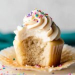 A Vanilla Cupcakes with a swirl of buttercream frosting and colorful sprinkles, with a bite taken out, showing its moist interior.