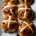 Freshly baked Vegan Hot Cross Buns with a shiny golden-brown crust, marked with a white cross and topped with dried raisins, arranged neatly on parchment paper.