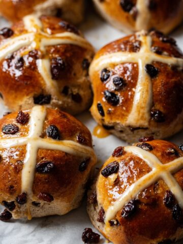 Easy Vegan Hot Cross Buns Recipe 1 Freshly baked Vegan Hot Cross Buns with a shiny golden-brown crust, marked with a white cross and topped with dried raisins, arranged neatly on parchment paper.