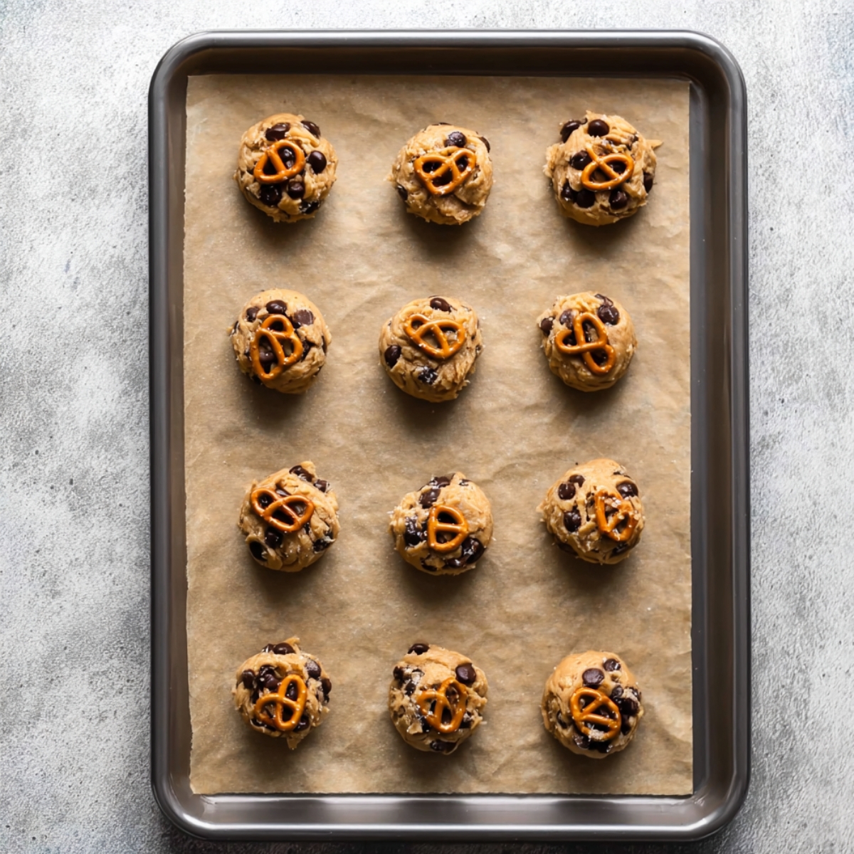 Kitchen Sink Cookies Recipe 11 A baking tray with twelve raw cookie dough balls, topped with pretzels and chocolate chips, ready to be baked.