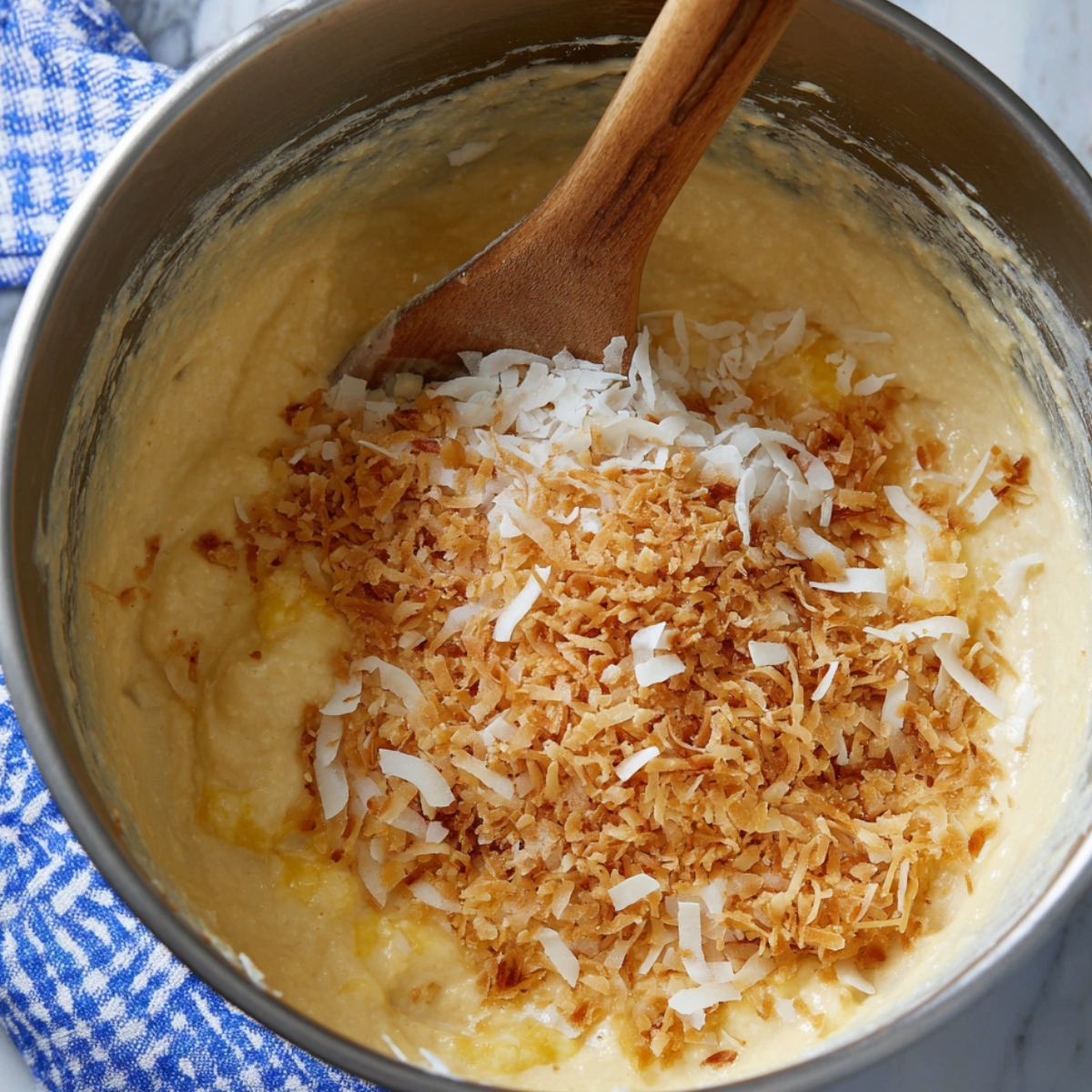 Easy Pineapple Coconut Bread Recipe 10 A metal mixing bowl with cake batter, shredded toasted coconut, and crushed pineapple being stirred with a wooden spoon.