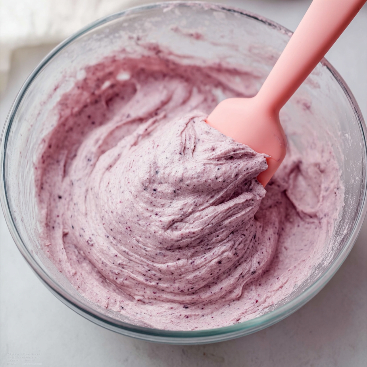 Easy Lemon Blueberry Cookies Recipe 11 A creamy, smooth blueberry frosting in a glass bowl, with a pale purple color and visible blueberry specks. A pink spatula is used to mix the frosting, showing its thick, fluffy texture.