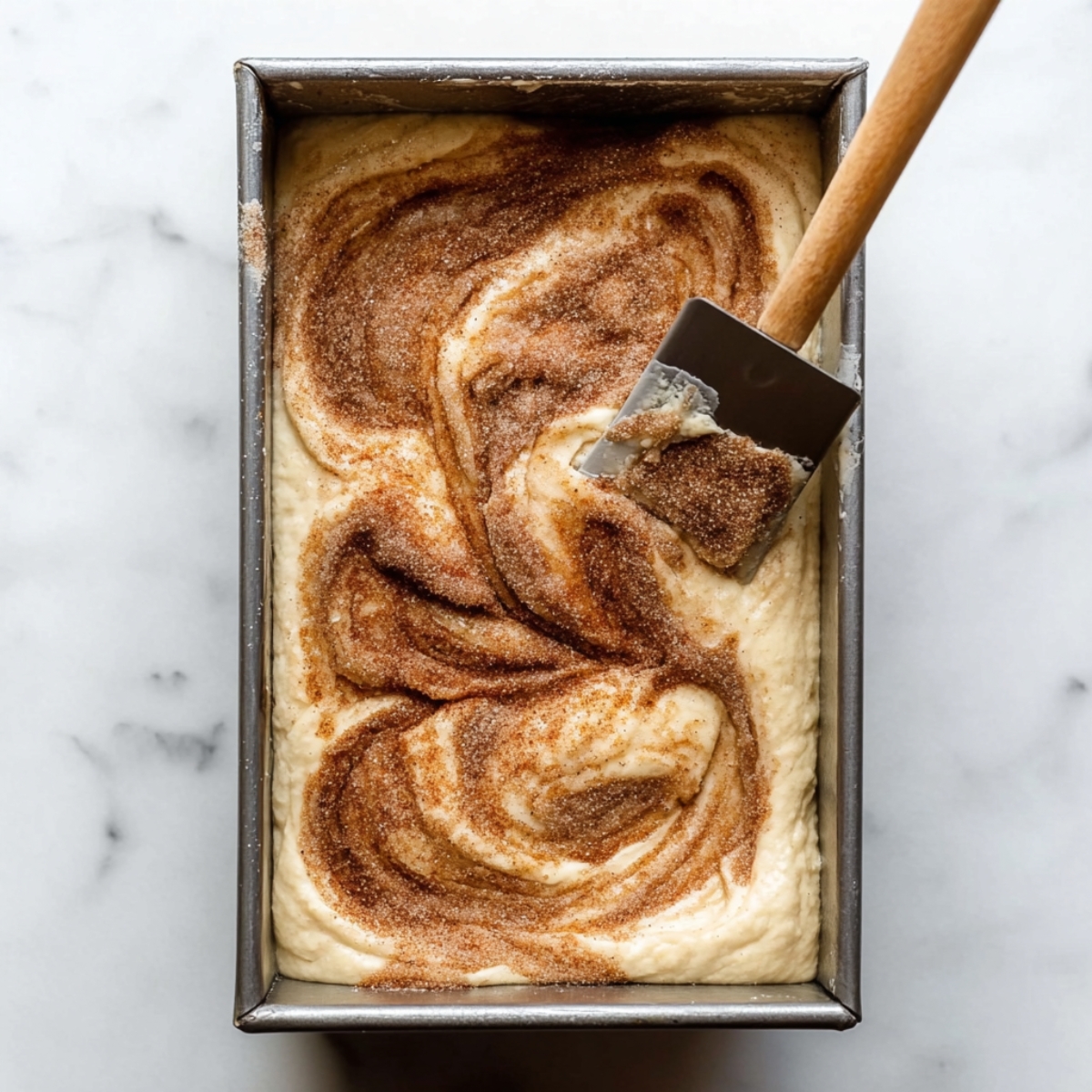 Delicious Cinnamon Swirl Quick Bread 11 A loaf pan filled with batter, topped with a swirl of cinnamon sugar. A spatula is gently swirling the cinnamon sugar into the batter, creating a marbled effect.