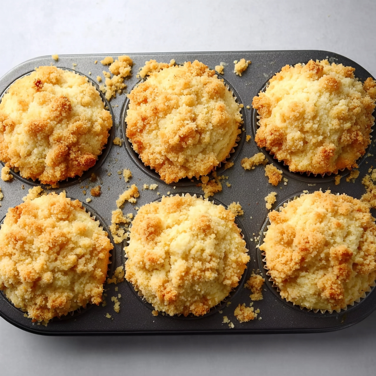 Lemon Curd Muffins with Streusel Topping 11 A muffin tin with six freshly baked lemon muffins, topped with a golden crumbly streusel topping. Some crumbs are scattered around the tin, showcasing the light and airy muffins.