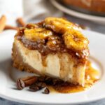 A slice of Bananas Foster Cheesecake with caramelized banana topping and syrup on a white plate.