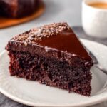 A slice of Chocolate Gooey Butter Cake with a rich, glossy ganache topping, served on a white plate, showcasing the moist interior.