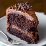 A slice of homemade Chocolate Layer Cake frosting, topped with mini chocolate chips, placed on a white plate with a fork beside it.