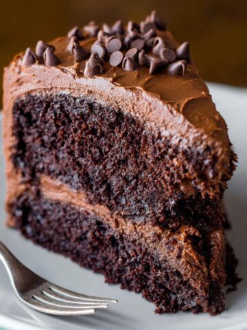 Deliciously Moist Chocolate Layer Cake 1 A slice of homemade Chocolate Layer Cake frosting, topped with mini chocolate chips, placed on a white plate with a fork beside it.