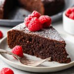 A slice of rich Chocolate Olive Oil Cake topped with fresh raspberries and a dusting of powdered sugar, served on a white plate.