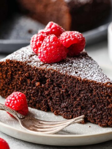 Chocolate Olive Oil Cake Recipe 1 A slice of rich Chocolate Olive Oil Cake topped with fresh raspberries and a dusting of powdered sugar, served on a white plate.
