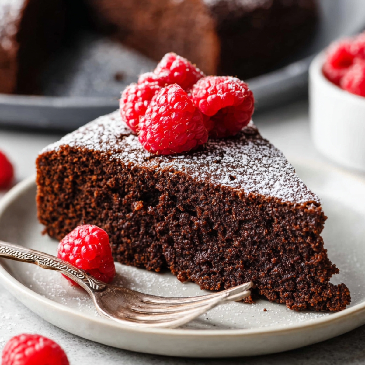 A slice of rich Chocolate Olive Oil Cake topped with fresh raspberries and a dusting of powdered sugar, served on a white plate.