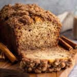 A freshly baked loaf of Cinnamon Streusel Banana Bread with a crumbly cinnamon streusel topping, sliced to reveal its soft, moist interior. The loaf is accompanied by cinnamon sticks and a cozy kitchen backdrop.