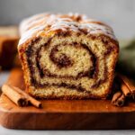 A close-up of a freshly baked Cinnamon Swirl Quick Bread, with a cinnamon sugar swirl inside, topped with a light drizzle of icing. The bread is placed on a wooden cutting board with cinnamon sticks.
