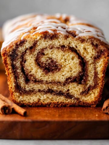 Delicious Cinnamon Swirl Quick Bread 1 A close-up of a freshly baked Cinnamon Swirl Quick Bread, with a cinnamon sugar swirl inside, topped with a light drizzle of icing. The bread is placed on a wooden cutting board with cinnamon sticks.