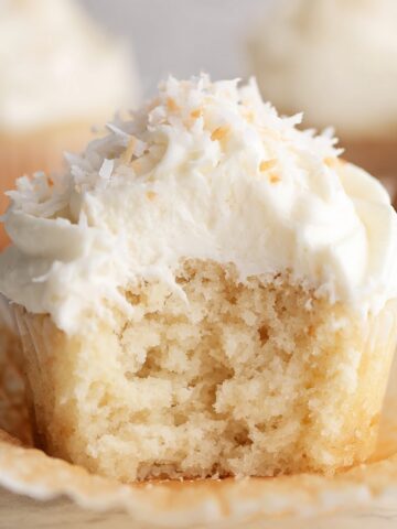 Easy Coconut Cupcakes Recipe 1 Fluffy Coconut Cupcakes with a bite taken out, topped with coconut flakes and creamy frosting.