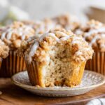 A close-up of a Coffee Cake Muffins with a crumbly streusel topping and a drizzle of icing, with a bite taken out, showing the fluffy interior of the muffin.