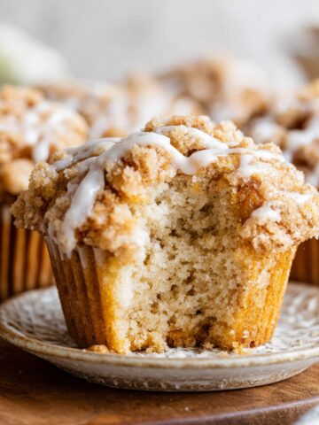 A close-up of a Coffee Cake Muffins with a crumbly streusel topping and a drizzle of icing, with a bite taken out, showing the fluffy interior of the muffin.