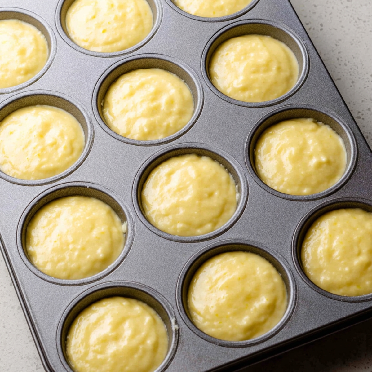 Healthy Lemon Muffins Recipe 11 A muffin tin filled with lemon muffin batter, evenly distributed in each of the twelve cups. The batter is smooth and slightly lumpy, ready to be baked into fluffy, golden muffins.