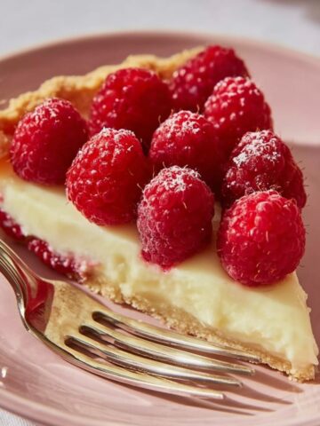 French Raspberry Tart Recipe 2 A slice of French Raspberry Tart topped with fresh raspberries, sitting on a pink plate. The tart has a creamy filling with a golden crust and is accompanied by a fork.