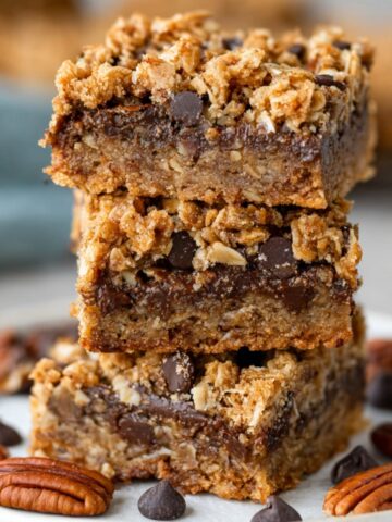 Gluten-Free Oatmeal Carmelita Bars 3 A stack of three Gluten-Free Oatmeal Carmelita Bars, showcasing the crumbly oat topping and gooey caramel and chocolate layers. The bars are surrounded by pecans and chocolate chips on a white plate.