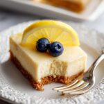 A slice of Greek Yogurt Lemon Bars on a plate, topped with a fresh lemon slice and blueberries, with a bite taken out to reveal the creamy filling.