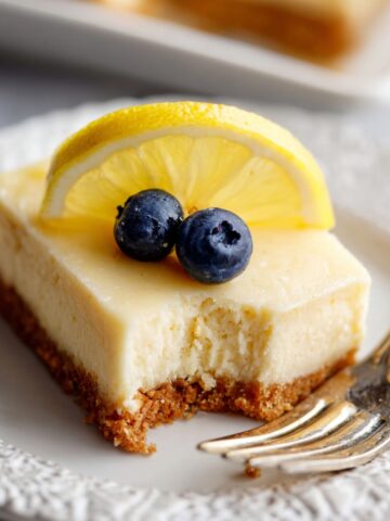Greek Yogurt Lemon Bars Recipe 1 A slice of Greek Yogurt Lemon Bars on a plate, topped with a fresh lemon slice and blueberries, with a bite taken out to reveal the creamy filling.