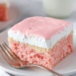 A slice of pink Hawaiian Guava Cake topped with white whipped cream and a smooth, pastel pink glaze, served on a white plate with a silver fork beside it.