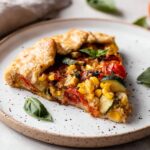 A slice of Homemade Savory Galette served on a white plate, featuring roasted tomatoes, zucchini, corn, and a golden, flaky crust with fresh basil on top.