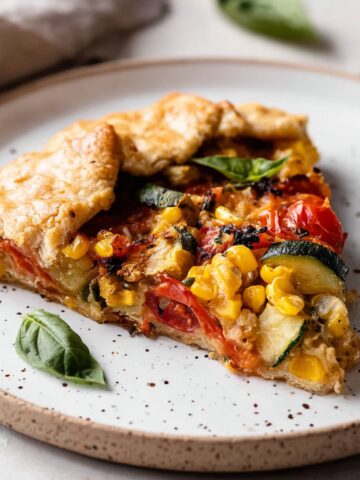 Easy Homemade Savory Galette Recipe 1 A slice of Homemade Savory Galette served on a white plate, featuring roasted tomatoes, zucchini, corn, and a golden, flaky crust with fresh basil on top.