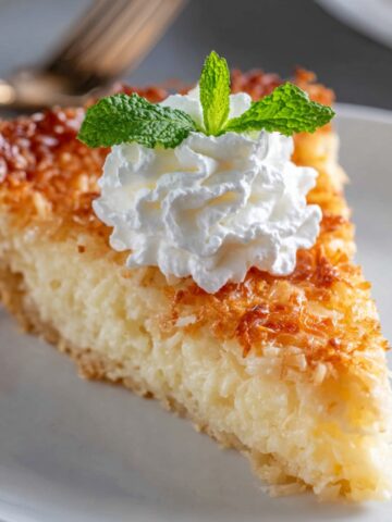 Easy Impossible Coconut Pie Recipe 1 A slice of golden Impossible Coconut Pie topped with whipped cream and a fresh mint leaf, served on a white plate.