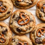 Close-up of freshly baked Kitchen Sink Cookies with pretzels, chocolate chips, and a sprinkle of sea salt on top, arranged on parchment paper.
