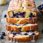 A loaf of Lemon Blueberry Bread, sliced to reveal moist crumb and juicy blueberries. The slices are drizzled with a sweet glaze, and scattered fresh blueberries are seen around the loaf.