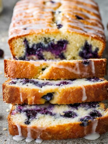 Easy Lemon Blueberry Bread Recipe 1 A loaf of Lemon Blueberry Bread, sliced to reveal moist crumb and juicy blueberries. The slices are drizzled with a sweet glaze, and scattered fresh blueberries are seen around the loaf.