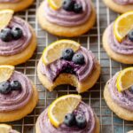 Soft Lemon Blueberry Cookies topped with blueberry frosting, a slice of lemon, and a blueberry. One cookie has a bite taken out, revealing the soft interior and the vibrant blueberry filling.