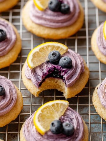 Soft Lemon Blueberry Cookies topped with blueberry frosting, a slice of lemon, and a blueberry. One cookie has a bite taken out, revealing the soft interior and the vibrant blueberry filling.