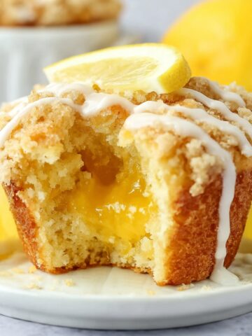 Lemon Curd Muffins with Streusel Topping 1 A Lemon Curd Muffins, with a crumbly topping, drizzled with icing and garnished with a slice of lemon. The muffin has been halved to reveal a gooey lemon curd center.