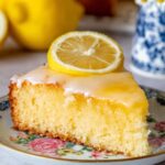 A slice of Lemon Gooey Butter Cake topped with lemon curd on a floral plate with whole lemons beside it.