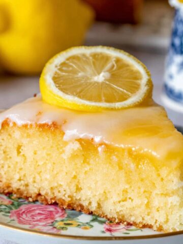 Delicious Lemon Gooey Butter Cake 2 A slice of Lemon Gooey Butter Cake topped with lemon curd on a floral plate with whole lemons beside it.