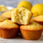 Freshly baked lemon muffins, one of them is cut in half to reveal the soft, fluffy interior. The muffins are golden brown with a moist and airy texture, and the bright lemon flavor is evident.