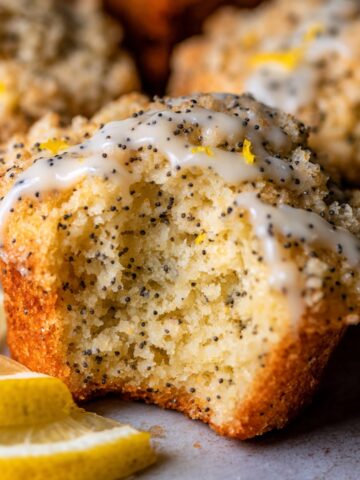 Moist Lemon Poppy Seed Muffins Recipe 1 A close-up of a Lemon Poppy Seed Muffins with a bite taken out, showcasing a fluffy interior, poppy seeds, and a crumbly topping with icing drizzle, accompanied by lemon slices.
